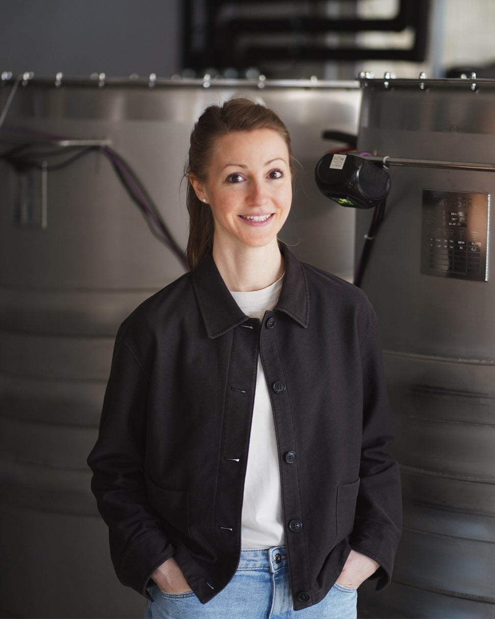 Die Gründerin von Erfrischerling, gekleidet mit einem weißen Thirt, einem schwarzen Hemd und einer hellblauen Jeans, die Hände lässig in die Hosentasche gesteckt, mit einem sympathischen Lächeln im Gesicht, vor 2 großen Edelstahltanks stehend